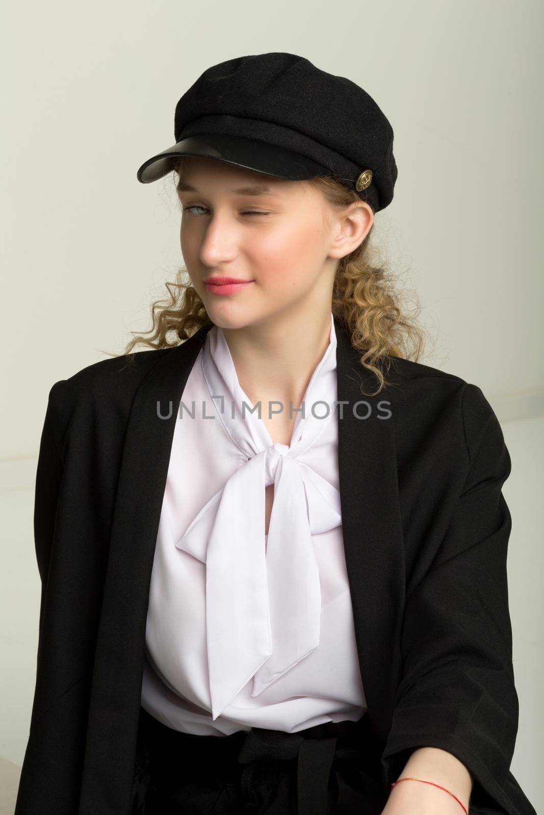 Portrait of elegant pretty girl blinking one eye. Beautiful stylish young woman in fashionable black blazer, white blouse and cap blinking at camera in playful manner against gray background in studio
