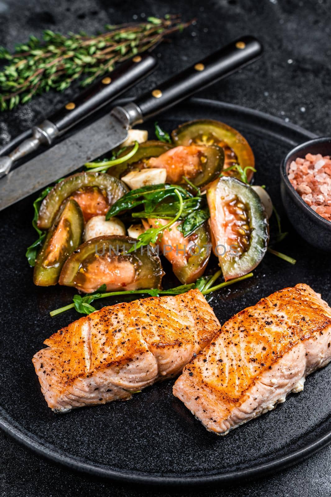 Fish meal with Roasted Salmon Fillet Steaks and arugula tomato salad on a plate. Black background. Top view.