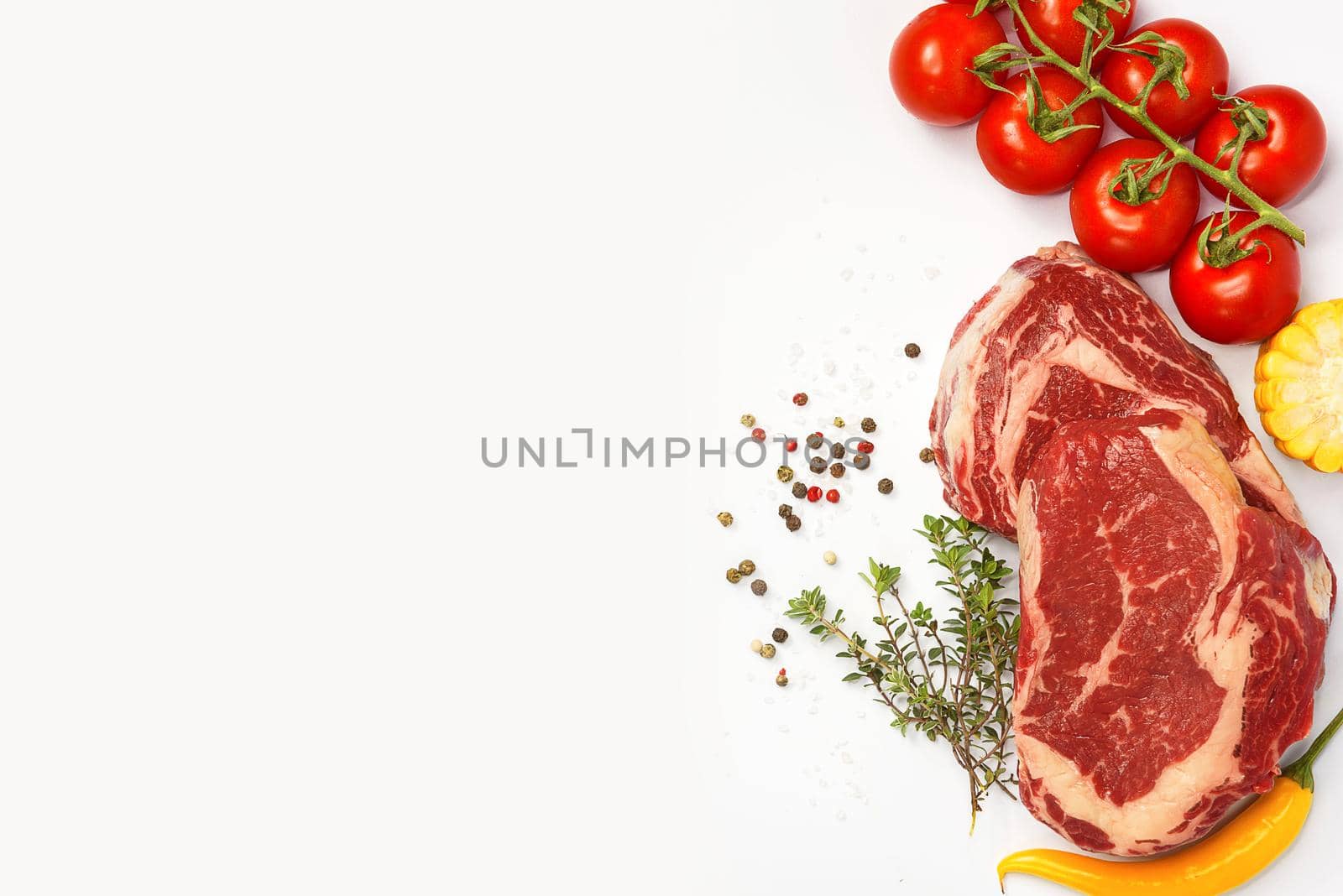 two fresh marbled beef steaks on a white plate with vegetables cherry tomatoes on a sprig, corn, spicy yellow pepper, a sprig of rosemary, salt and different varieties of peppers