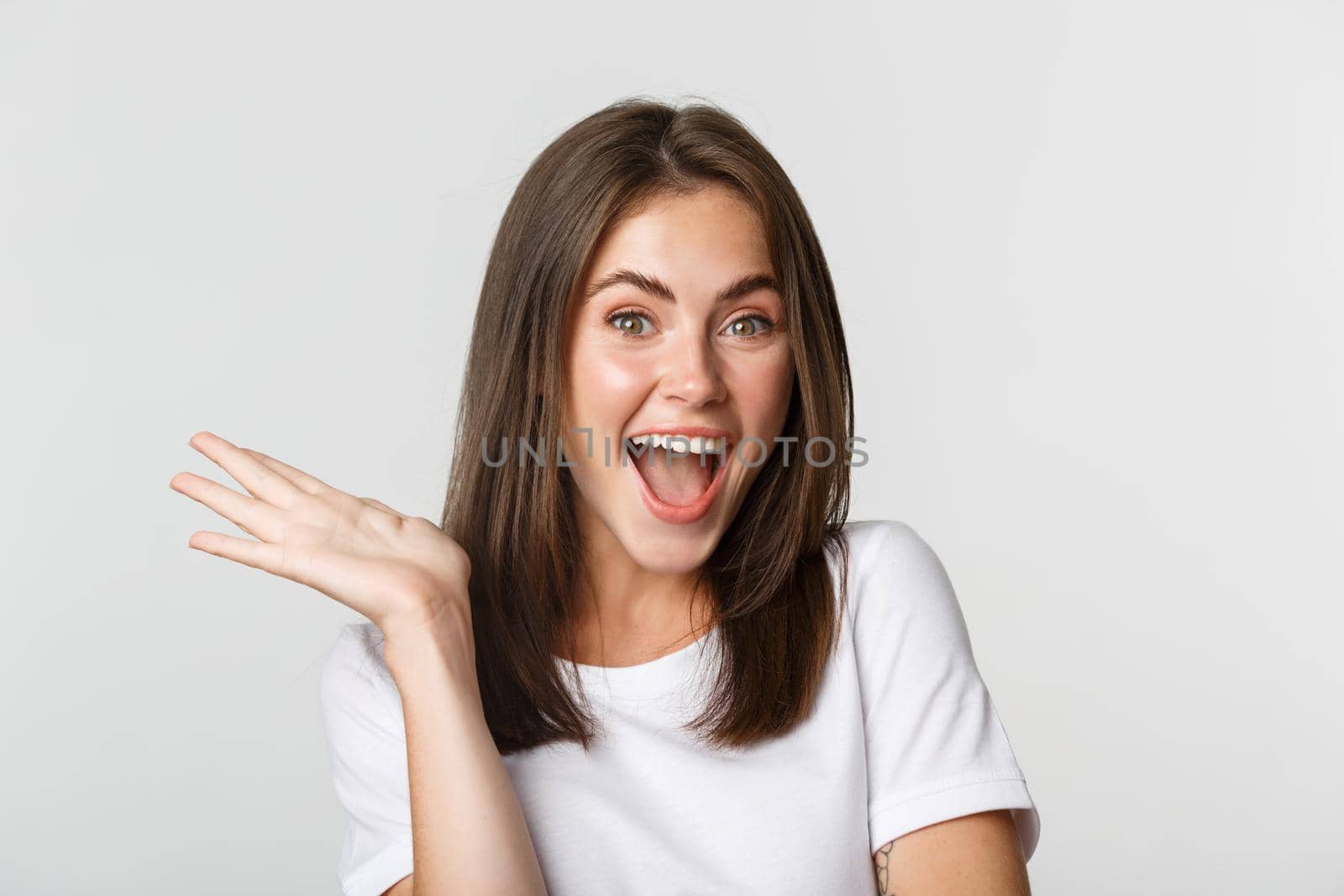 Close-up of excited beautiful brunette girl looking amused, waving hand to say hello, white background.