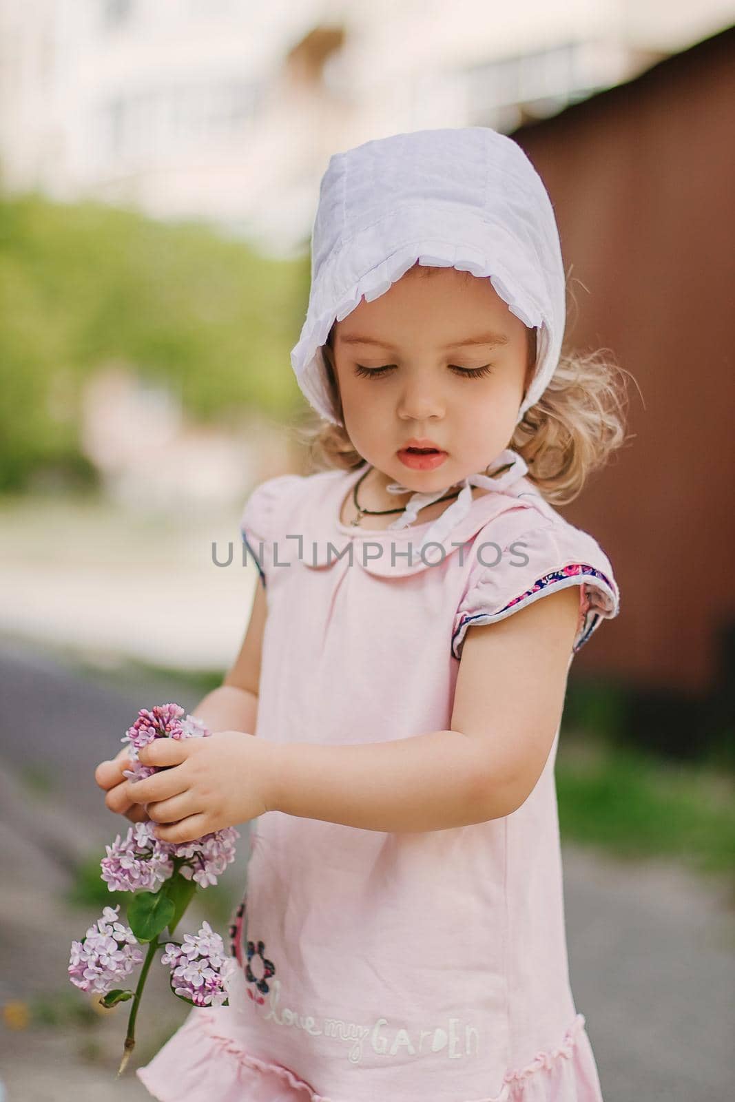 A beautiful little girl in a pink dress holds a branch of lilac flowers. Child in the garden on a background of a wooden fence with flowers in her hands.