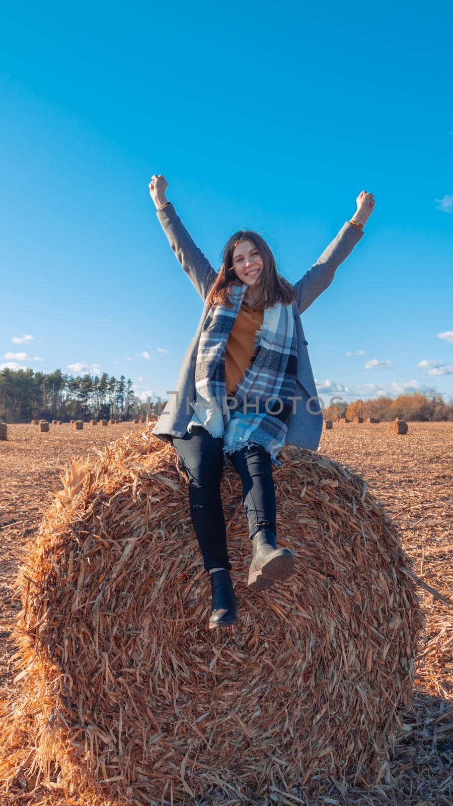 A girl of European appearance in a gray coat stands in a field near a larger bale with hay against a blue sky. by lunarts