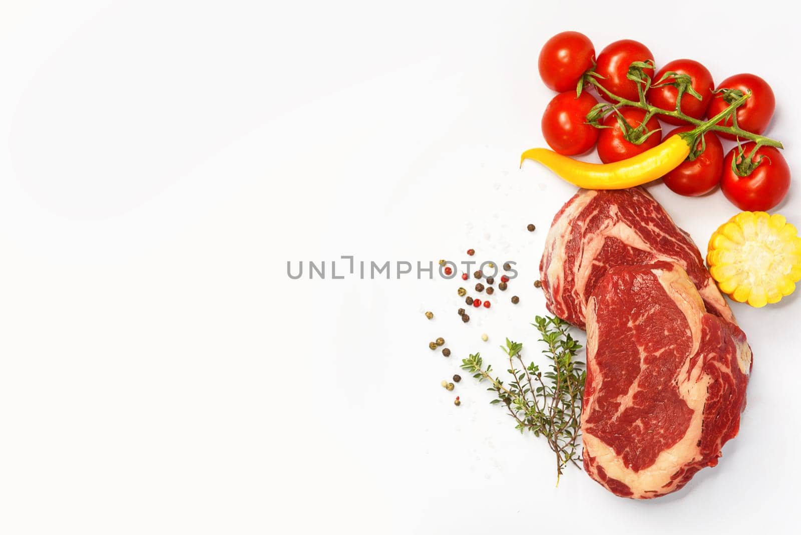 two fresh marbled beef steaks on a white plate with vegetables cherry tomatoes on a sprig, corn, spicy yellow pepper, a sprig of rosemary, salt and different varieties of peppers