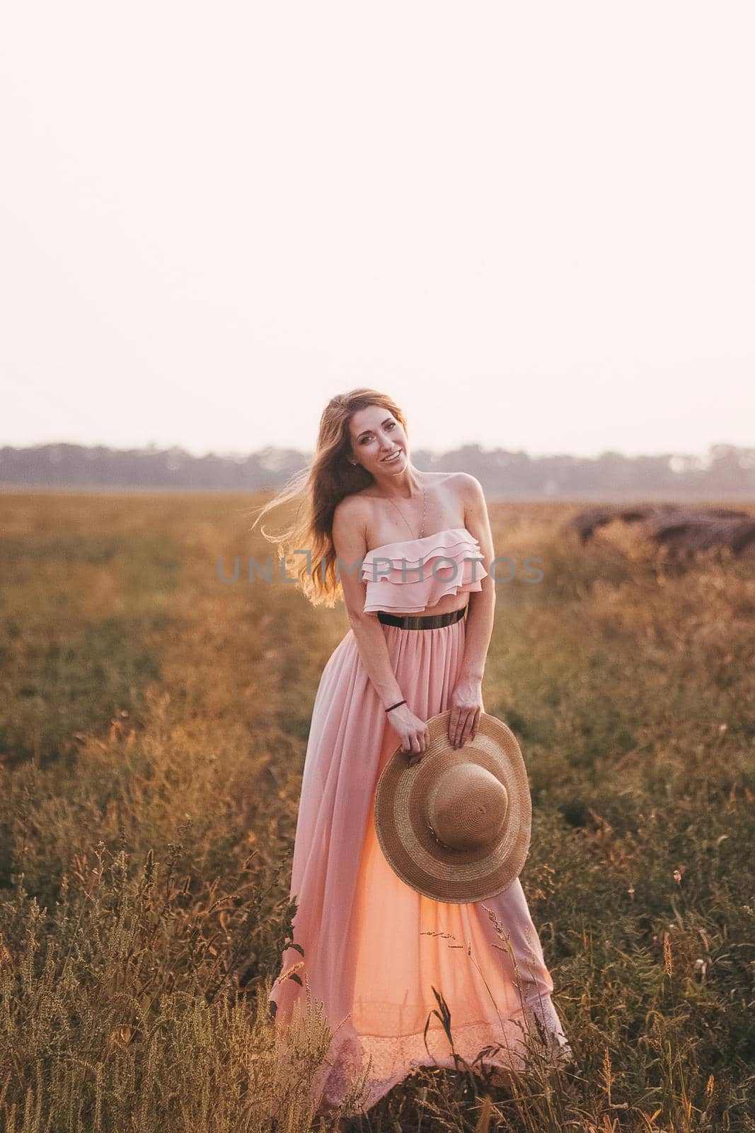 Beautiful romantic model girl outdoors dressed in tender long dress in the field in sunset light. Wind blowing long hair. Glow Sun, Sunshine. Backlit. Toned in warm colors.
