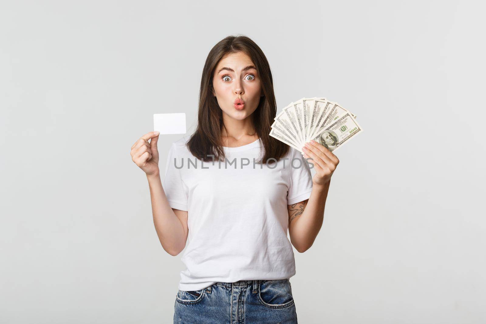 Amused brunette girl looking excited, holding money and credit card, standing white background.