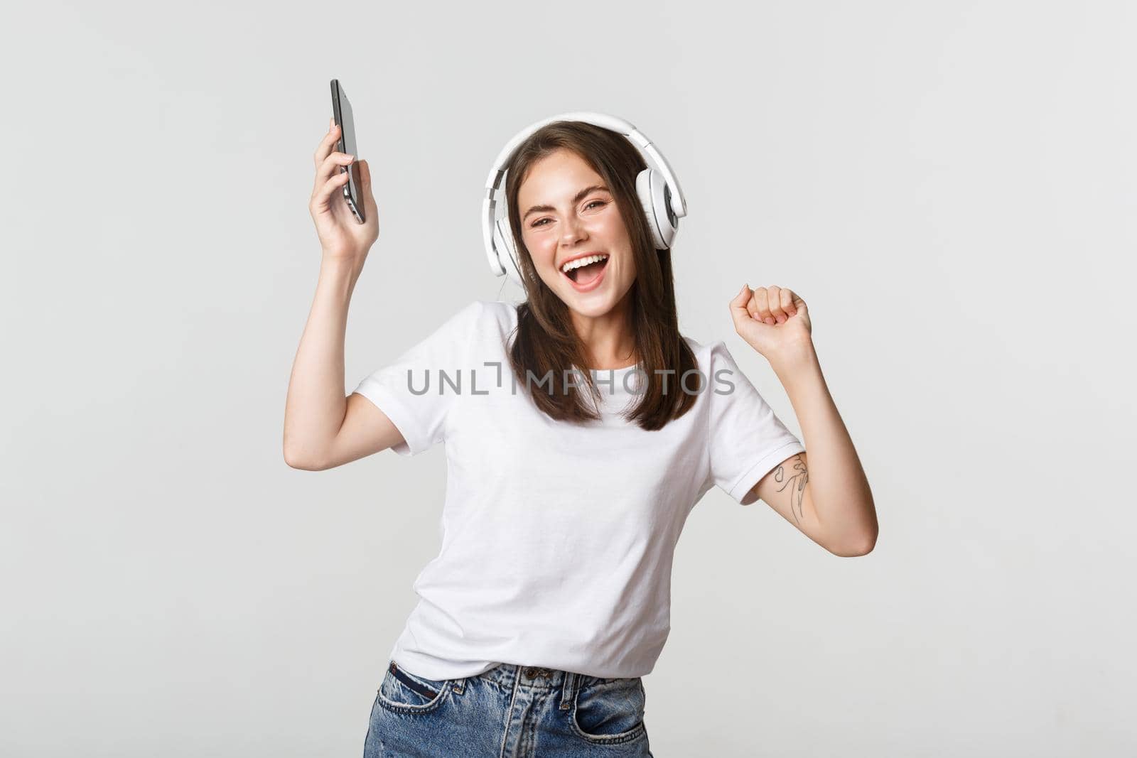 Happy beautiful brunette girl dancing and listening music in wireless headphones, holding smartphone.