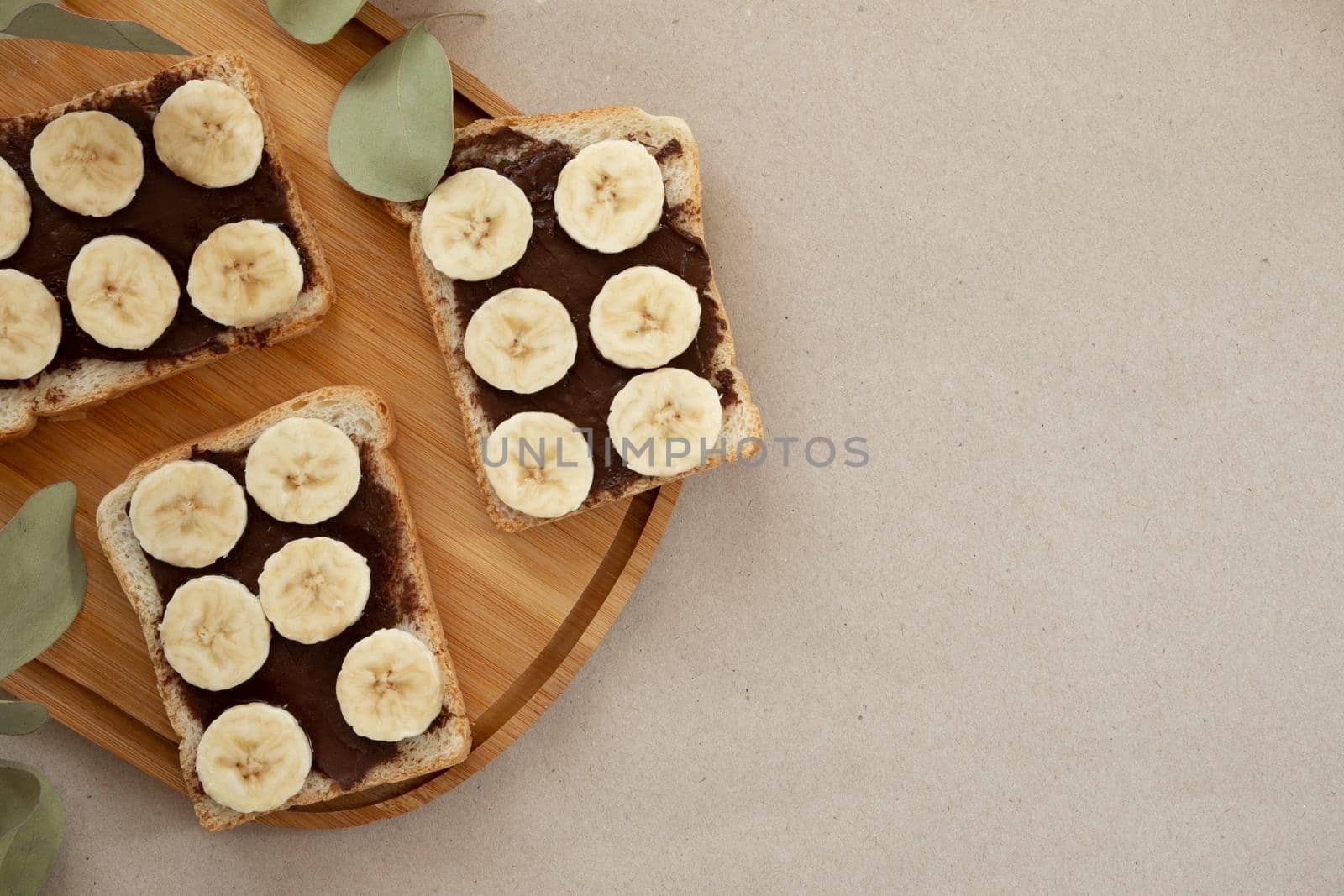 Three banana white bread toasts smeared with chocolate butter that lie on a cutting board with a sprig of leaves on craft paper background. top view with area for text