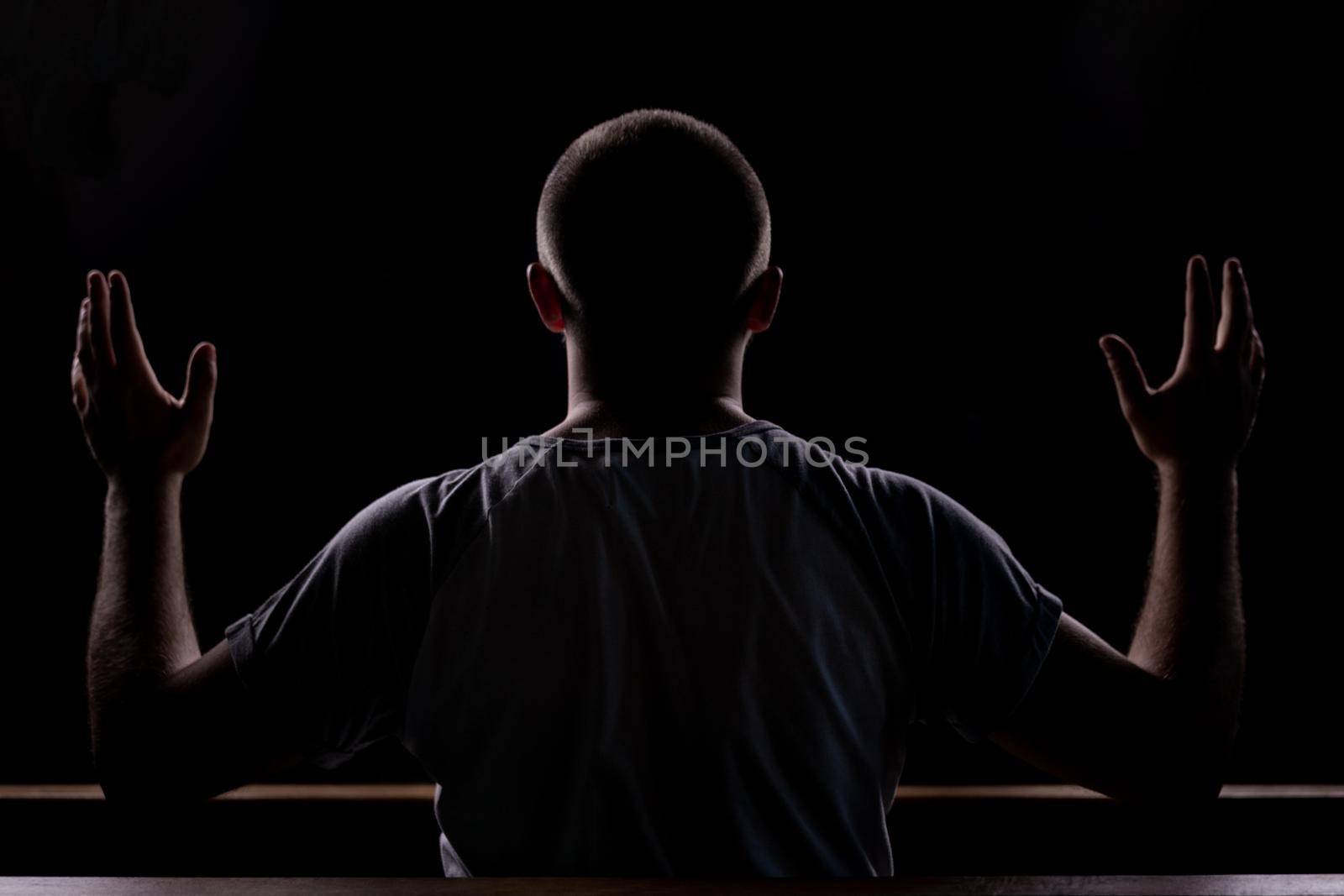 Silhouette of a young guy who sits in church and praying with hands raised up. Close-up view from behind. Backlight.