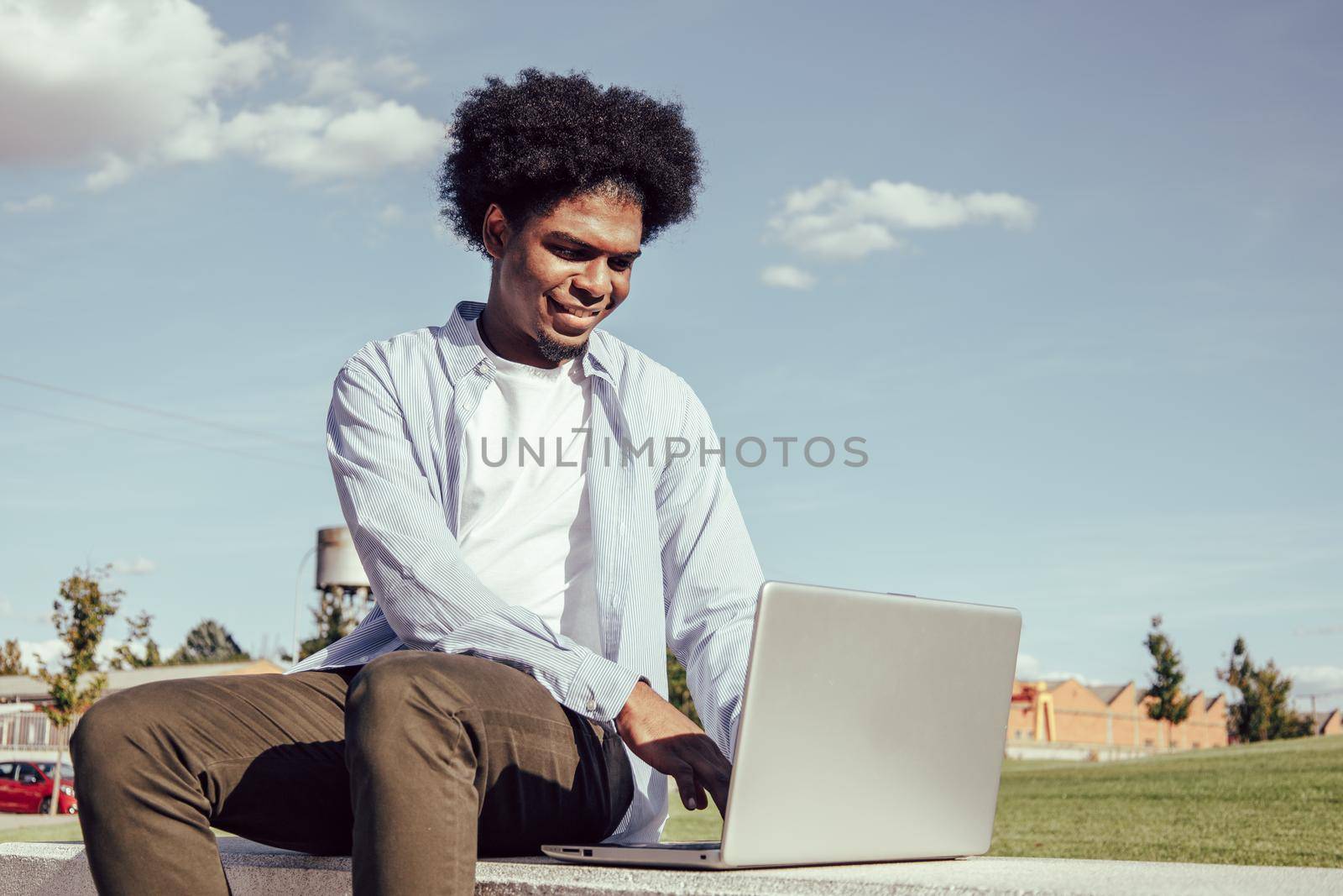 Young African American Guy Working On Laptop Outside, Sitting On Bench In Park. by ALVANPH