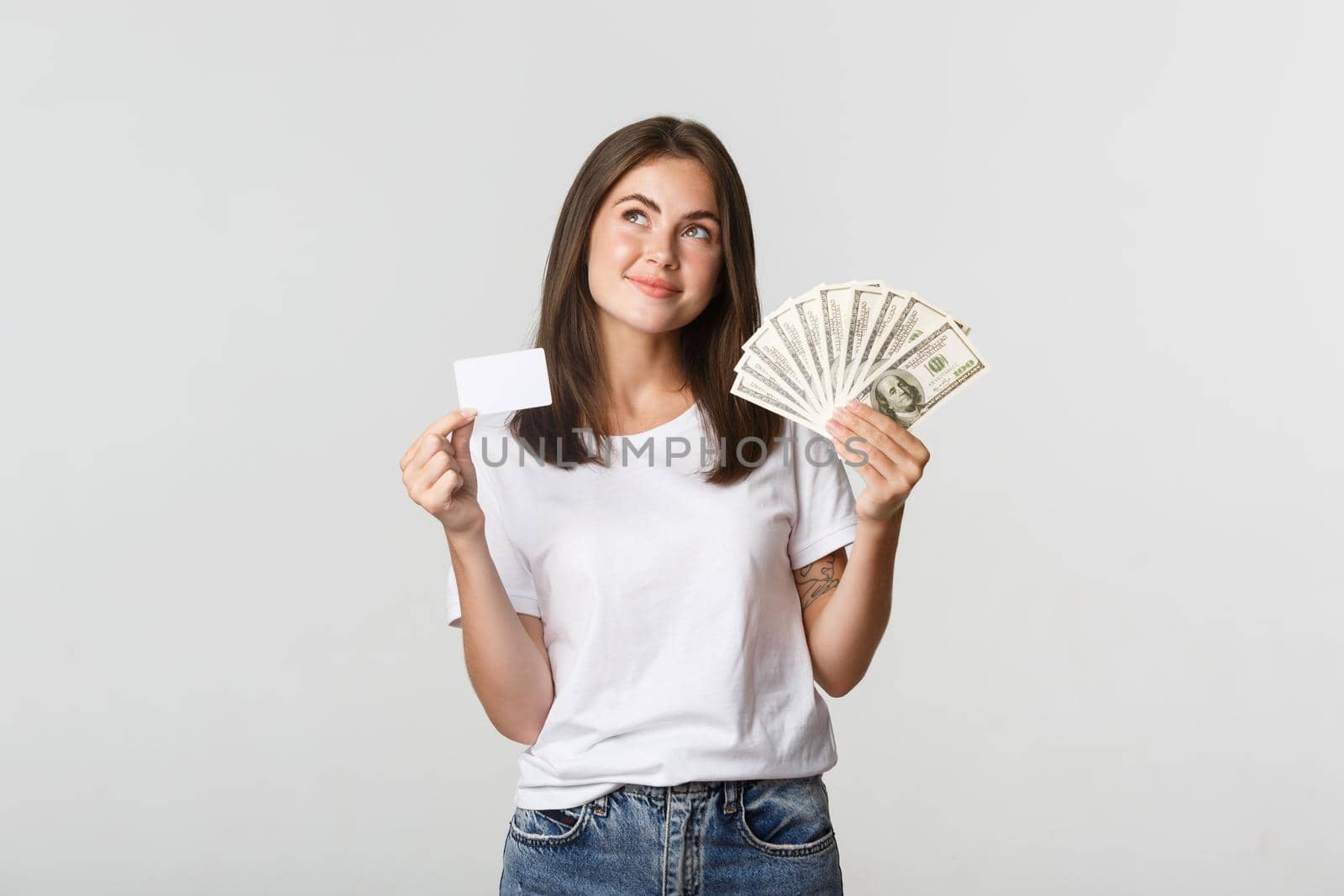 Thoughtful smiling girl holding money and credit card, looking upper left corner, standing white background and pondering.