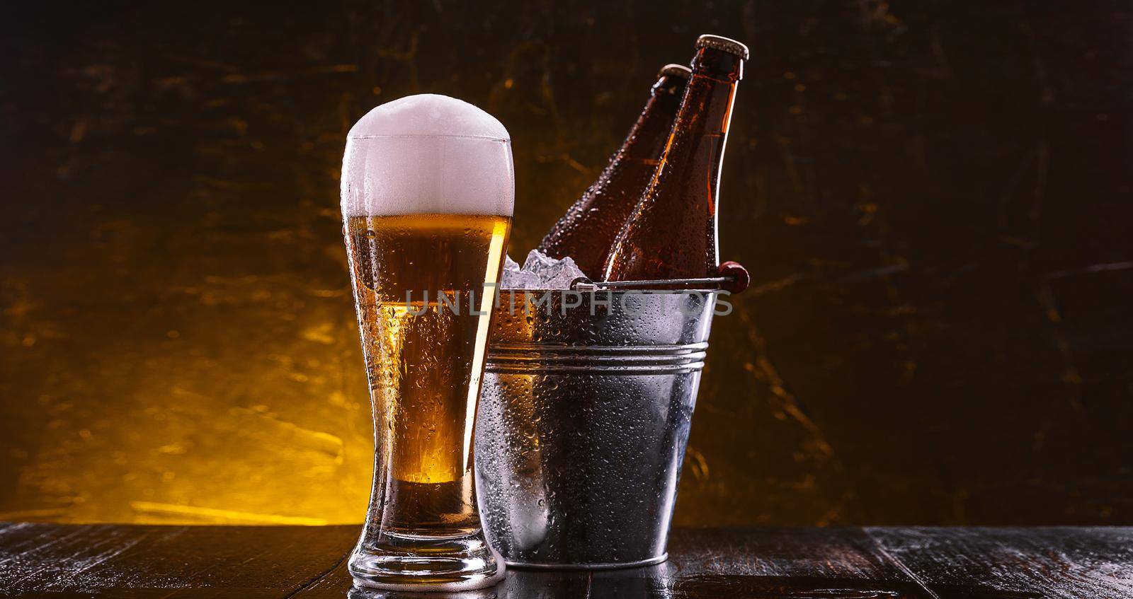two bottles of beer in a bucket with ice and a glass of beer with lush foam next to a dark background
