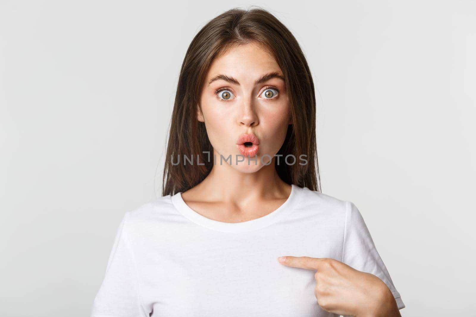 Close-up of surprised and excited young brunette girl pointing at herself, white background.