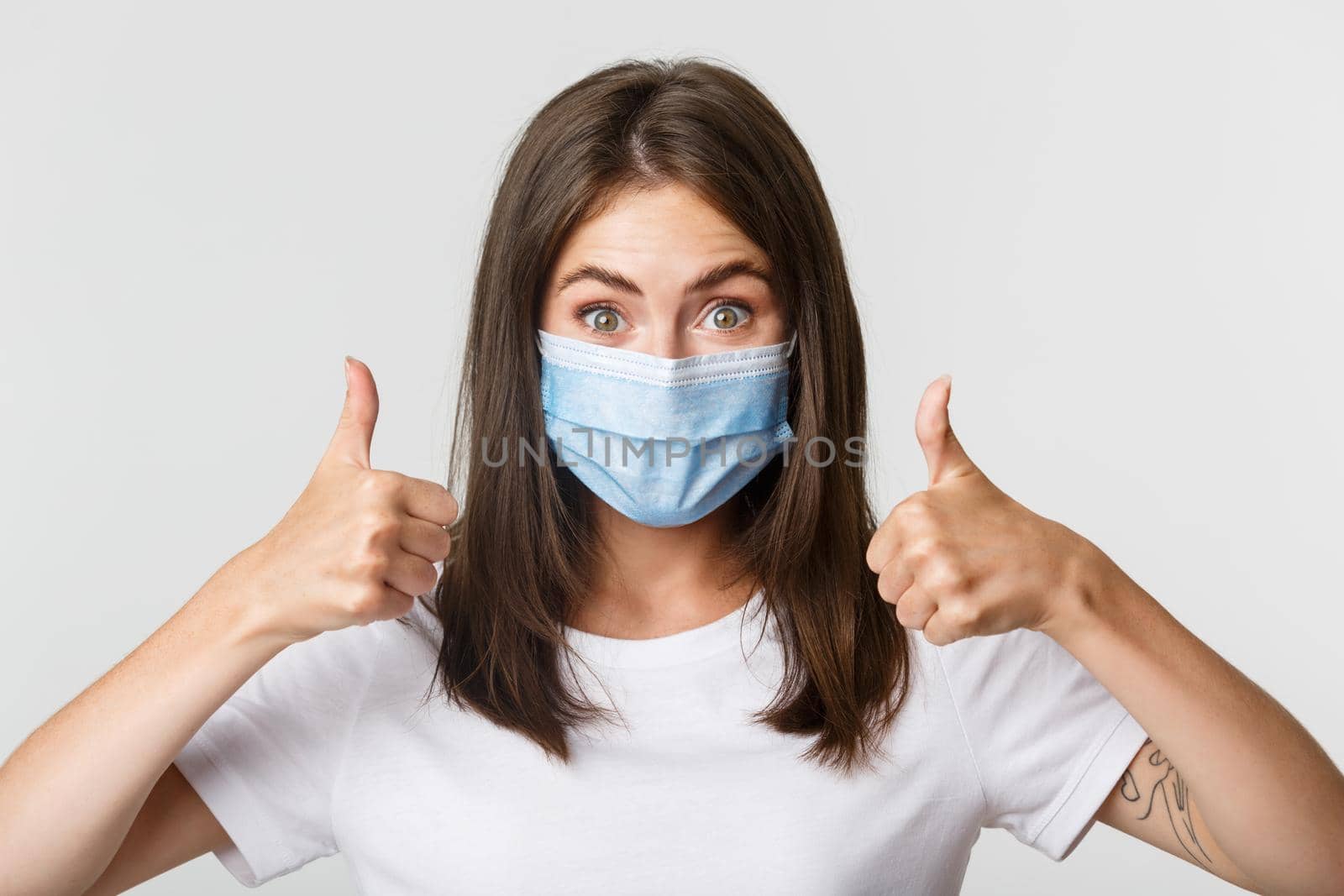 Covid-19, health and social distancing concept. Close-up of pleased attractive girl in medical mask showing thumbs-up in approval.