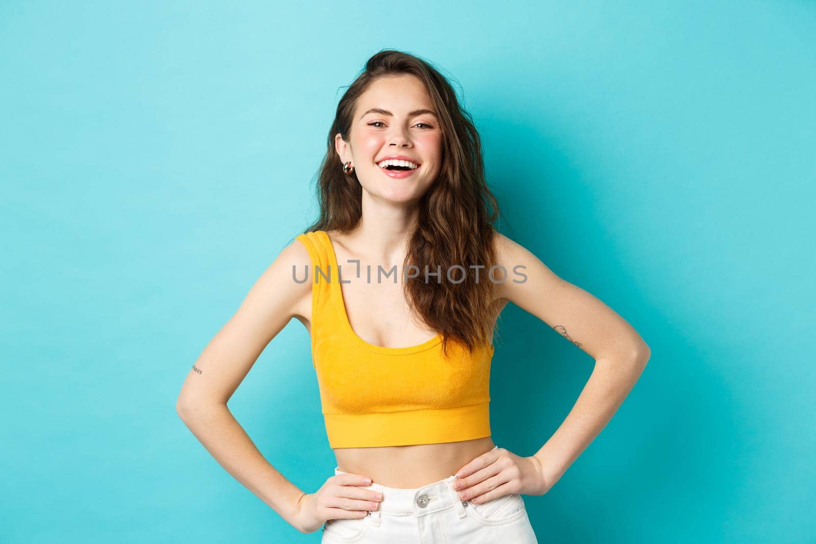 Image of confident and happy stylish woman laughing, holding hands on waist and smiling cheerful, standing carefree in summer outfit, blue background by Benzoix