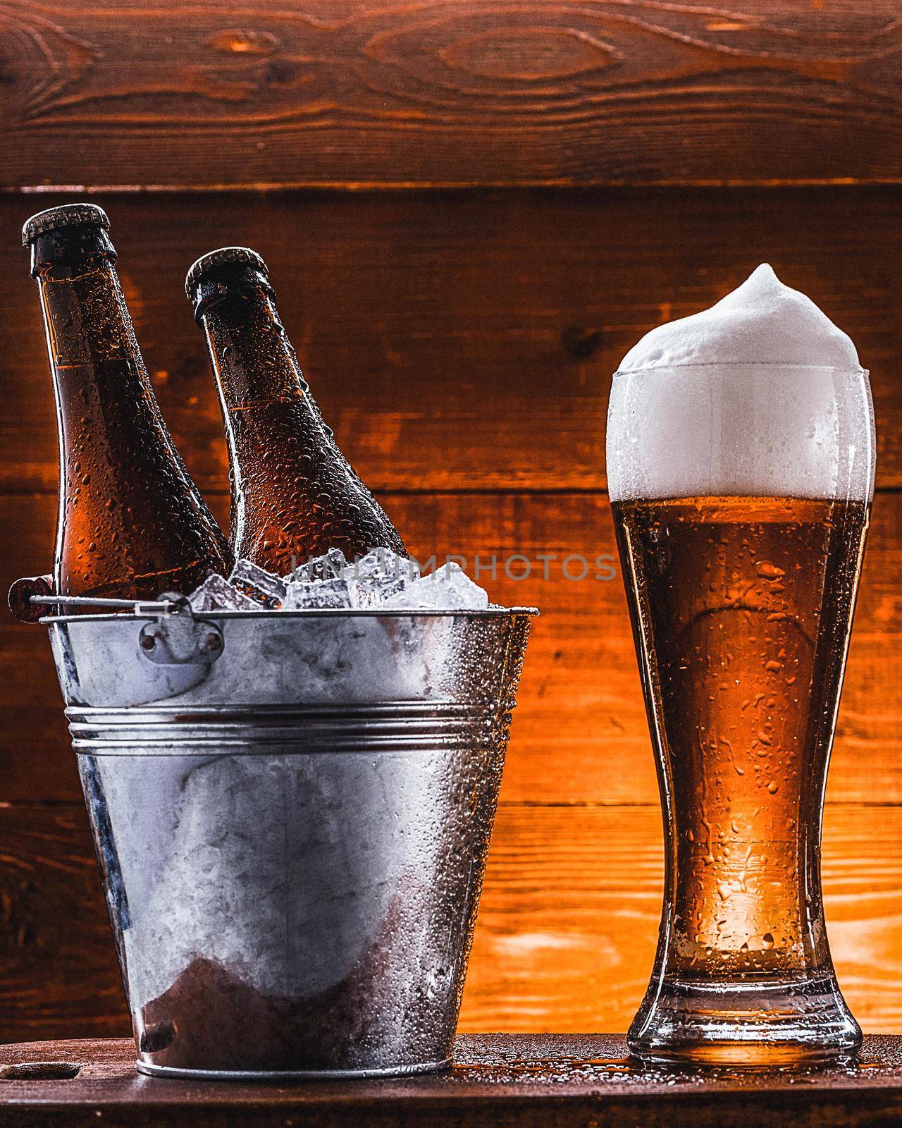 two bottles of beer in a bucket with ice and a glass of beer with lush foam next to a dark background by vvmich