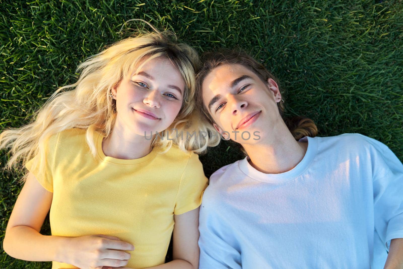 Happy couple of teenagers on green grass. Close-up of smiling faces of guy and girl 16, 17 years old, top view. Youth, adolescence, happiness, relationship, lifestyle, young people concept