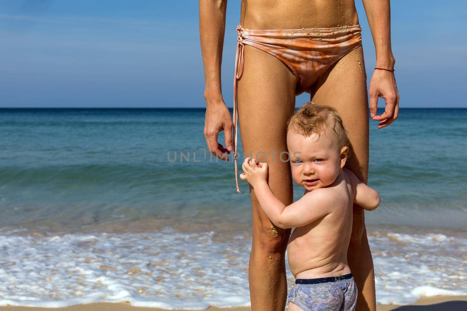 Cute healthy little baby boy in panties wet after sea bath looking at camera over shoulder while standing with mother in bikini on sunny beach