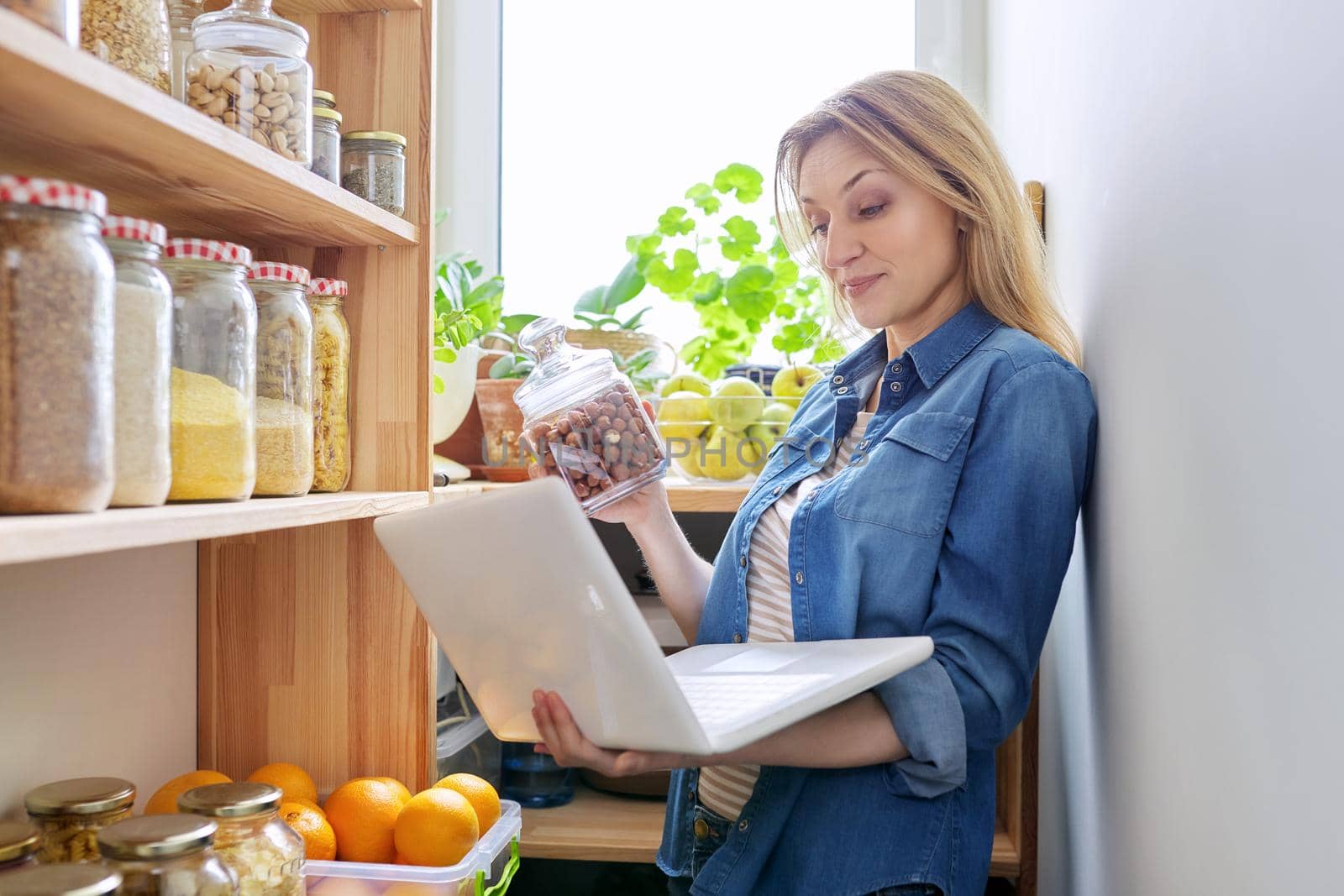 Middle-aged woman in kitchen in pantry with laptop showing cans of food. Female food blogger, online communication, healthy eating video recording, healthy food, technology, hobbies and leisure