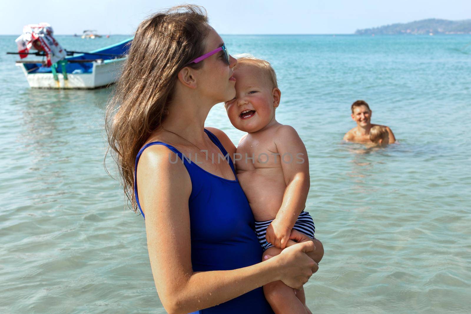 Young mom in swimsuit with playful child with open mouth on hands standing in water swimming in sea on sunny day looking at camera