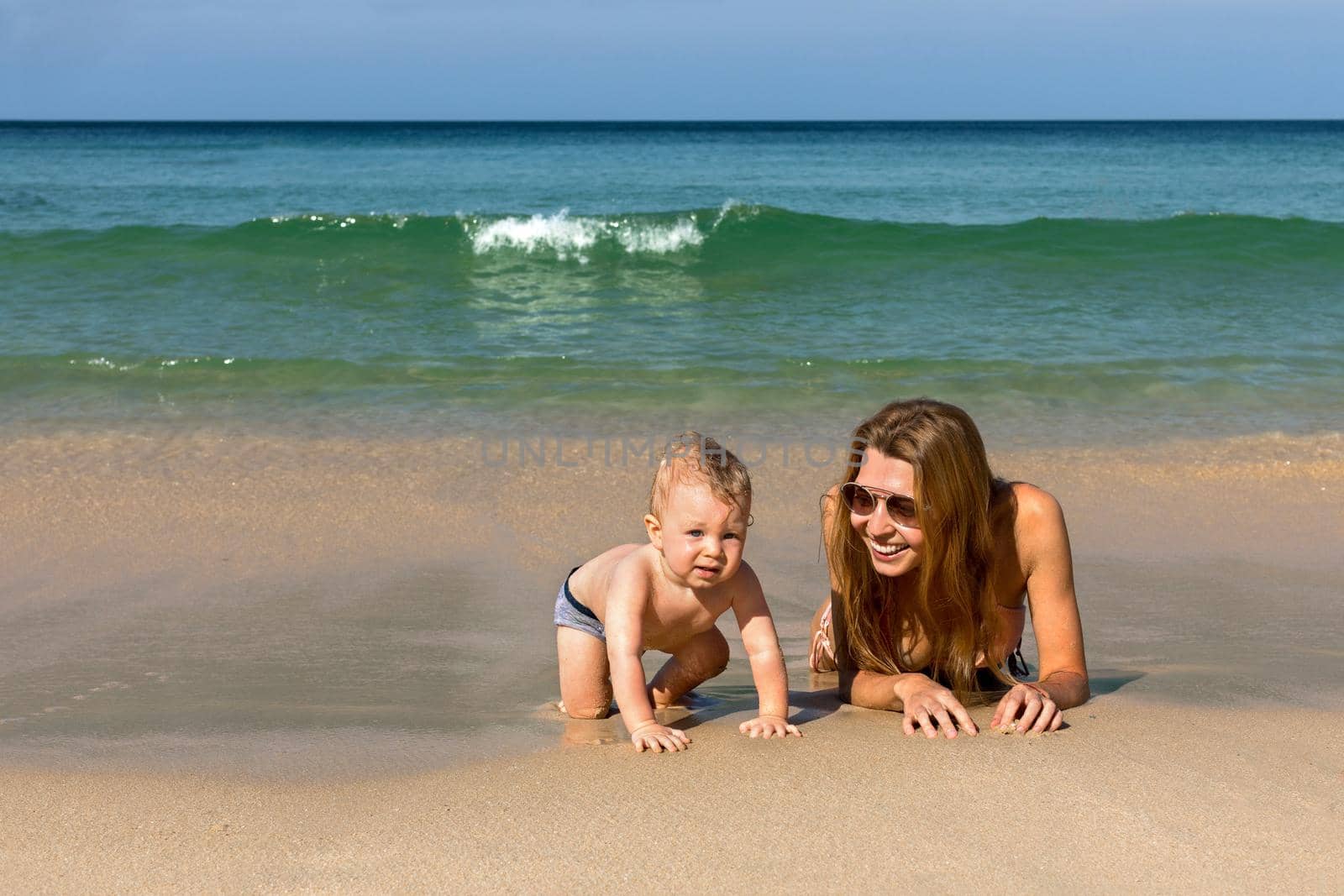 Cheerful young woman in bikini and sunglasses lying with crawling infant baby on sandy beach next to water and enjoying summer day and happy moments together