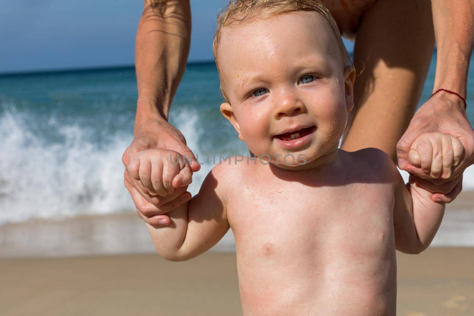 Adorable healthy little undressed baby with wet hair looking at camera while standing with help of crop mother on sandy beach in sunny day