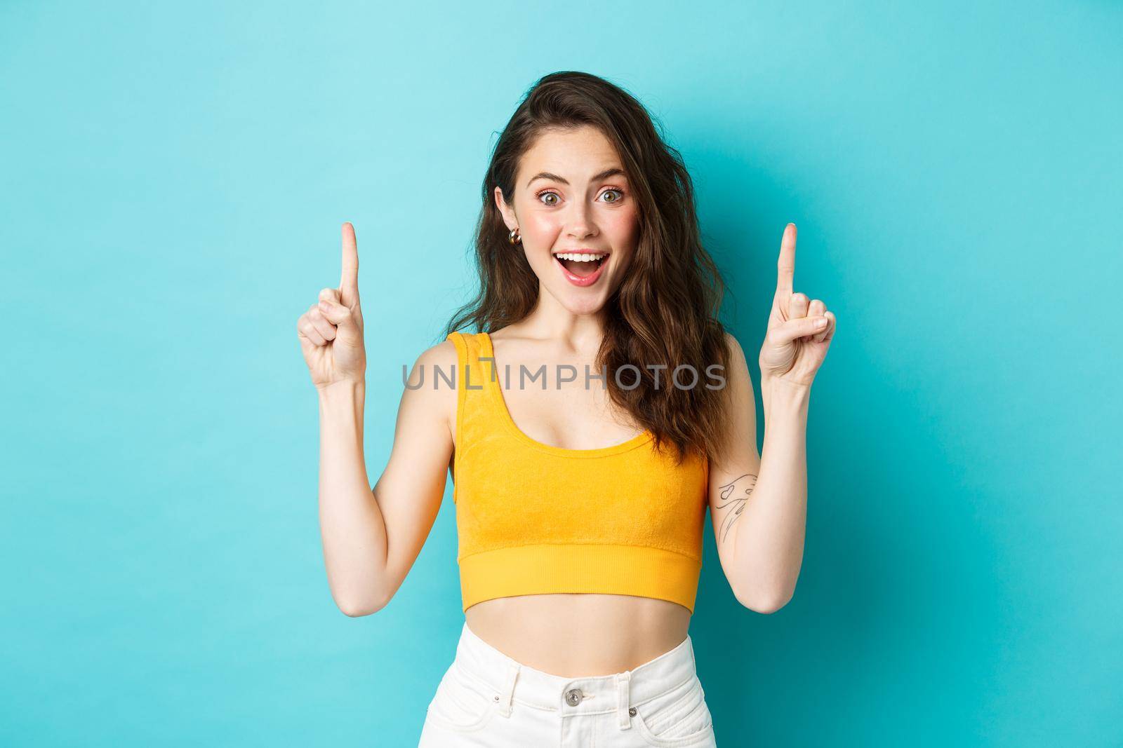 Summer lifestyle concept. Surprised happy woman showing advertisement on top, pointing fingers up and smiling excited, checking out special deal, standing over blue background.