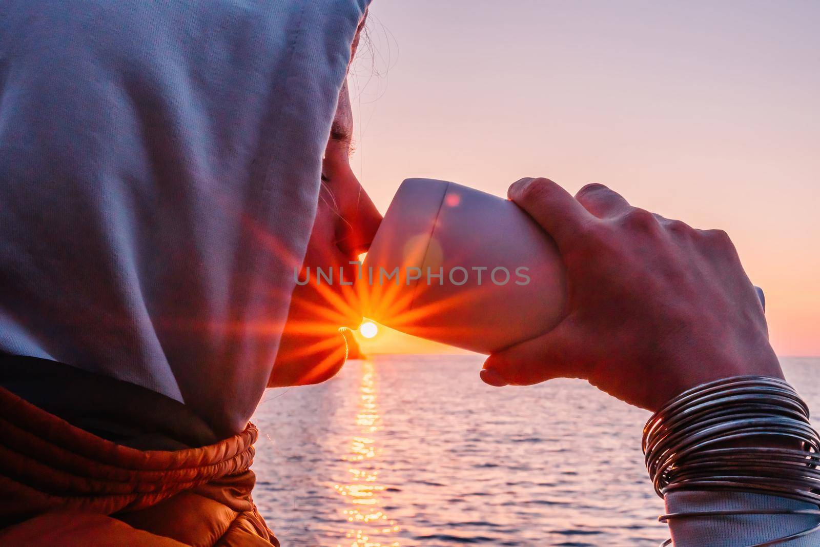 A young tourist Woman holding coffee tumbler cup while sitting outdoor and enjoying sunrise over sea mountain landscape. Women's yoga fitness routine. Healthy lifestyle, harmony and meditation by panophotograph