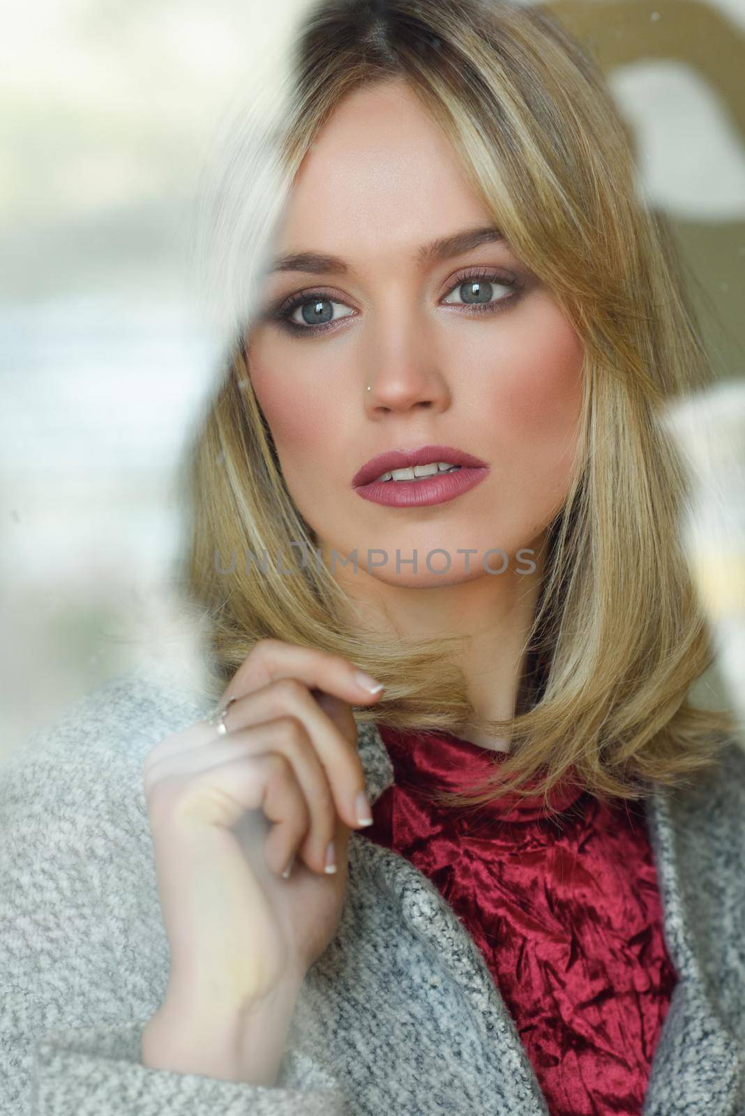 Thoughtful young blonde woman with lost eyes from the window. Beautiful girl at home wearing winter clothes. Pretty female with straight hair and blue eyes.