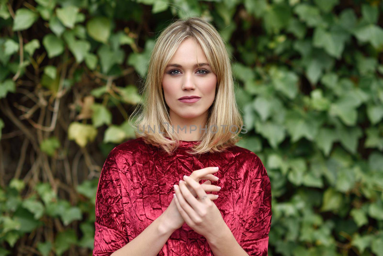 Beautiful blonde woman in green leaves background. Young girl wearing red dress standing in the street. Pretty female with straight hairstyle and blue eyes.