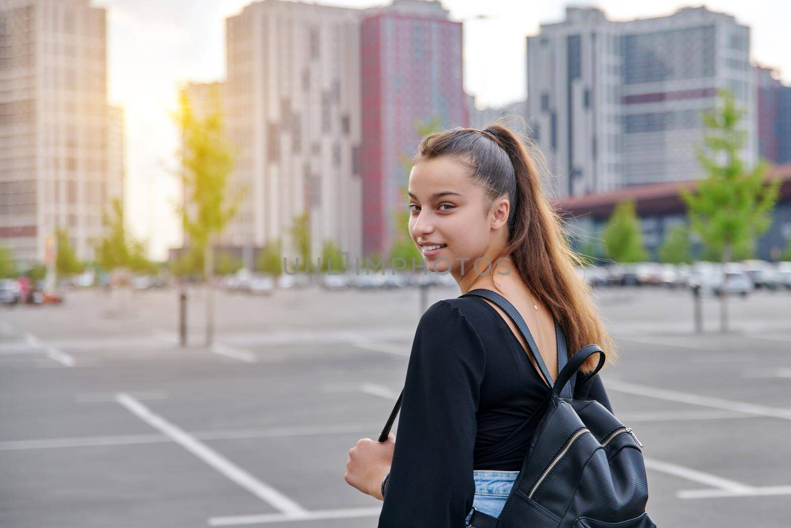 Fashionable beautiful smiling teenage girl in shorts with backpack looking at camera in big city parking lot, urban style, fashion, beauty, adolescence concept