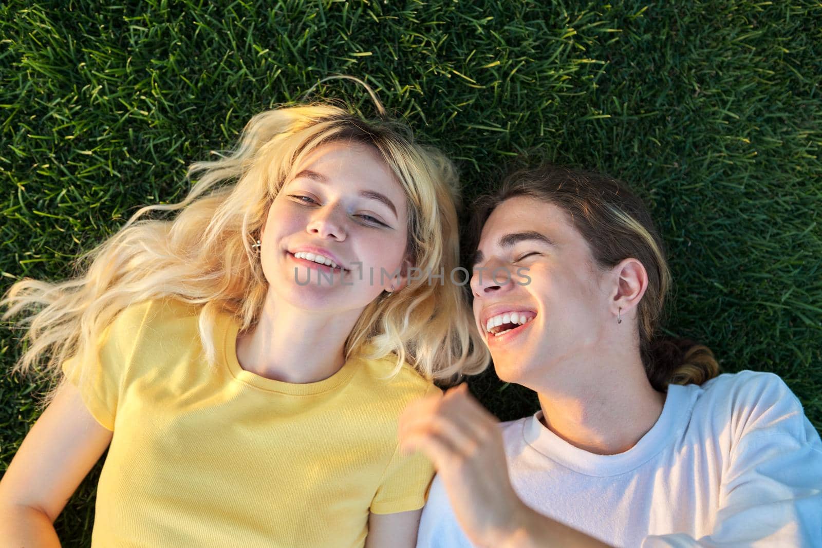 Happy couple of teenagers on green grass. Close-up of smiling faces of guy and girl 16, 17 years old, top view. Youth, adolescence, happiness, relationship, lifestyle, young people concept