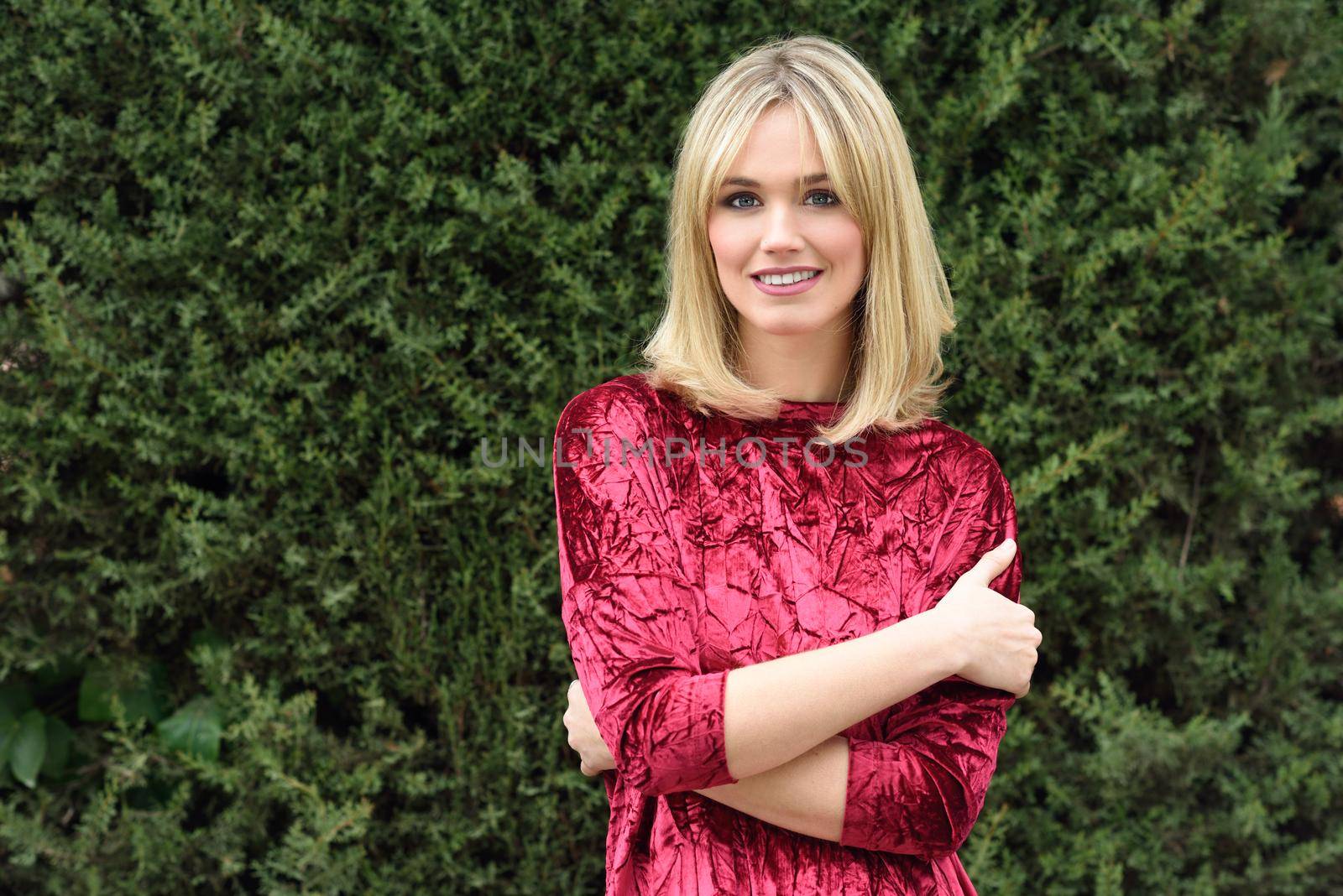 Beautiful blonde woman in green leaves background. Young girl wearing red dress standing in the street. Pretty female with frizzy hairstyle and blue eyes.