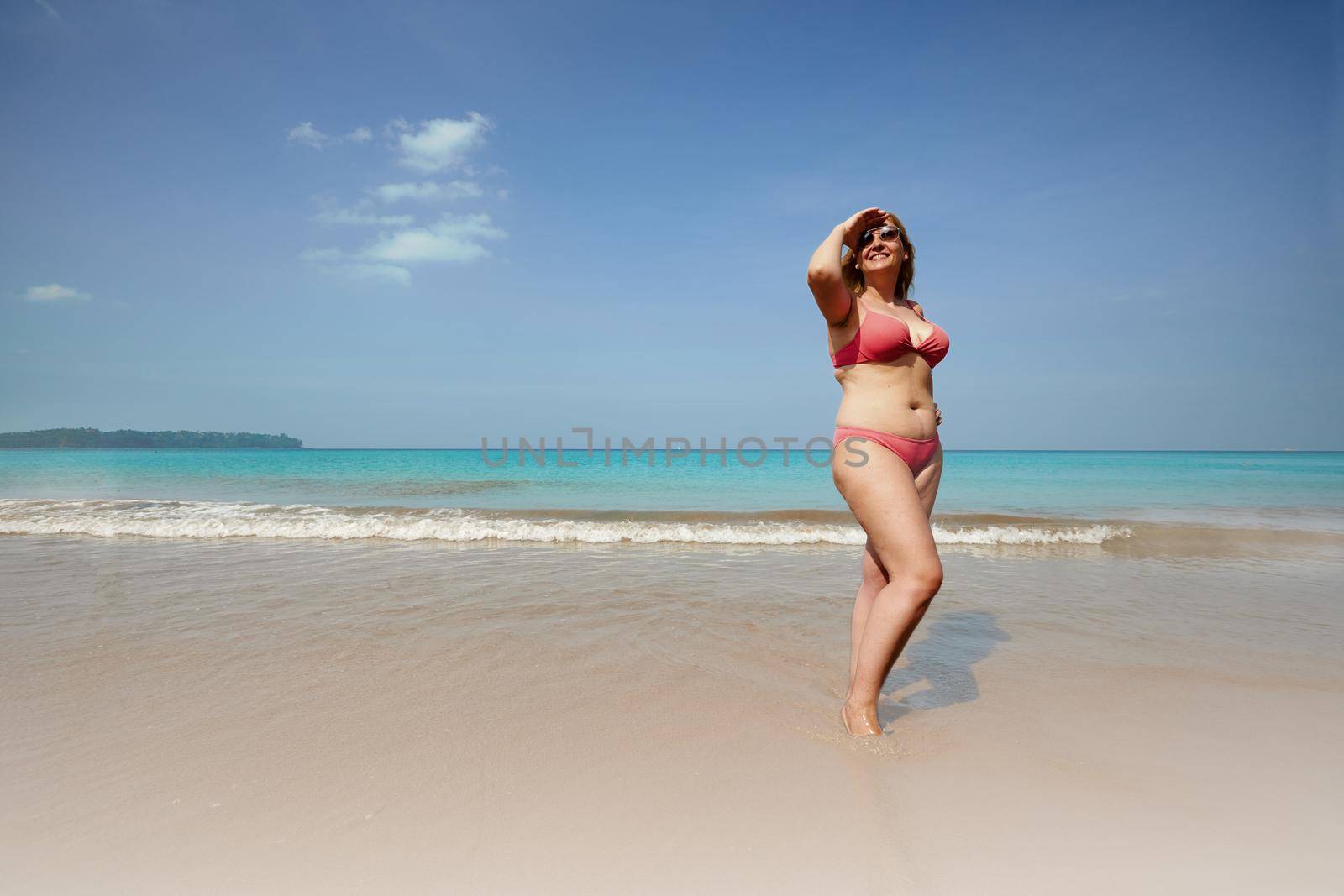 Full body of smiling overweight woman covering face from sunlight while standing on sandy beach near waving sea