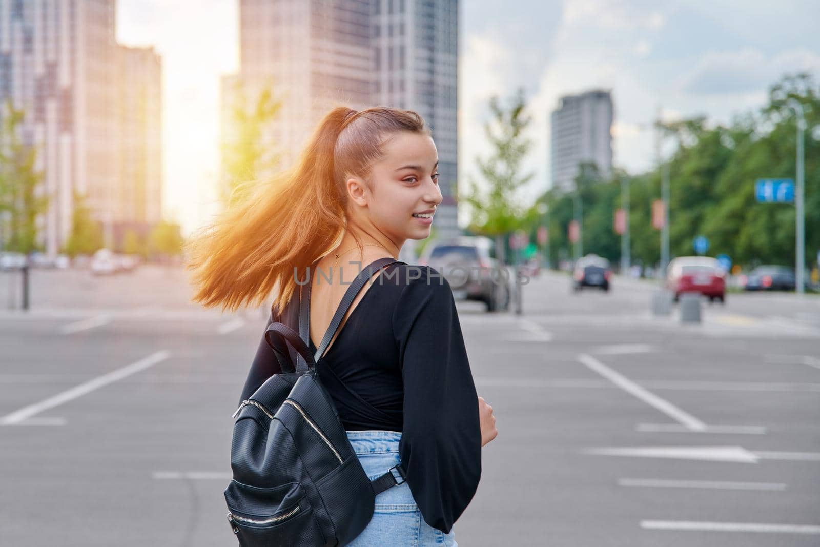 Fashionable beautiful smiling teenage girl in shorts with a backpack in a big city parking lot, urban style, fashion, beauty, adolescence concept