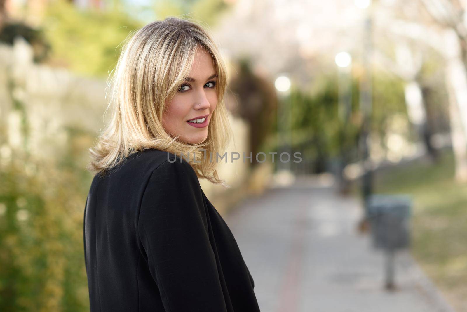 Beautiful blonde woman in urban background. Young girl wearing black blazer jacket standing in the street. Pretty female with straight hair hairstyle and blue eyes.