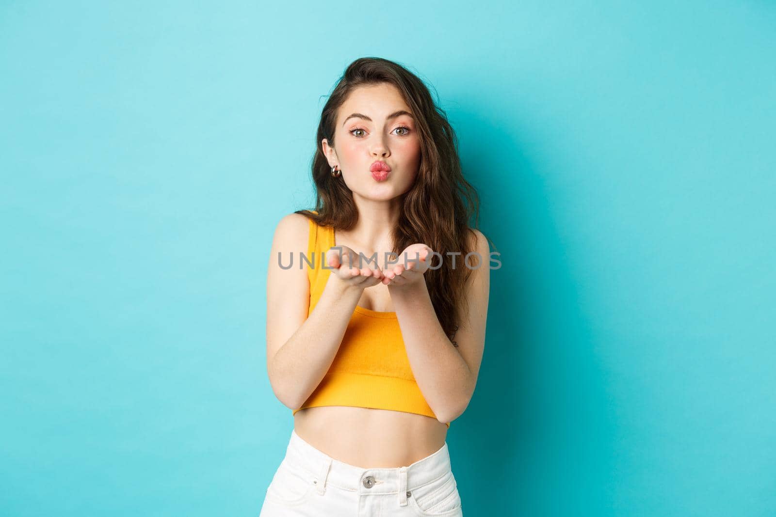 Beautiful glamour girl in summer clothes pucker her lips, blowing air kiss, sending mwah in palms, standing over blue background.