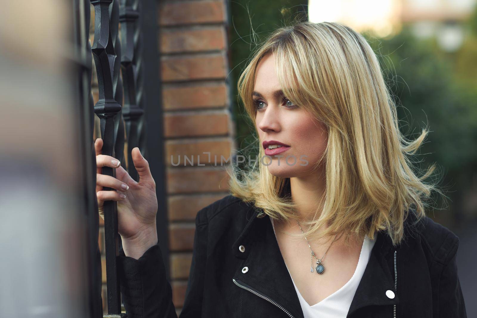 Attractive blonde woman standing in urban background with lost look. Young girl wearing black zipper jacket. Pretty female with straight hair hairstyle and blue eyes.