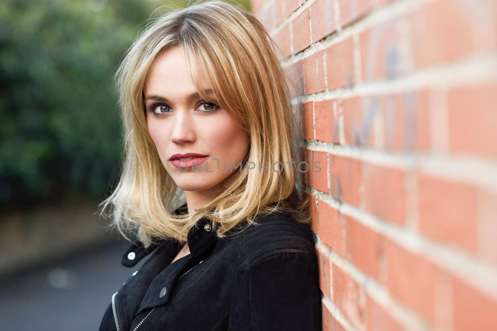 Pretty blonde woman in urban background. Young girl wearing black zipper jacket standing in the street. Pretty female with straight hair hairstyle and blue eyes on brick wall.