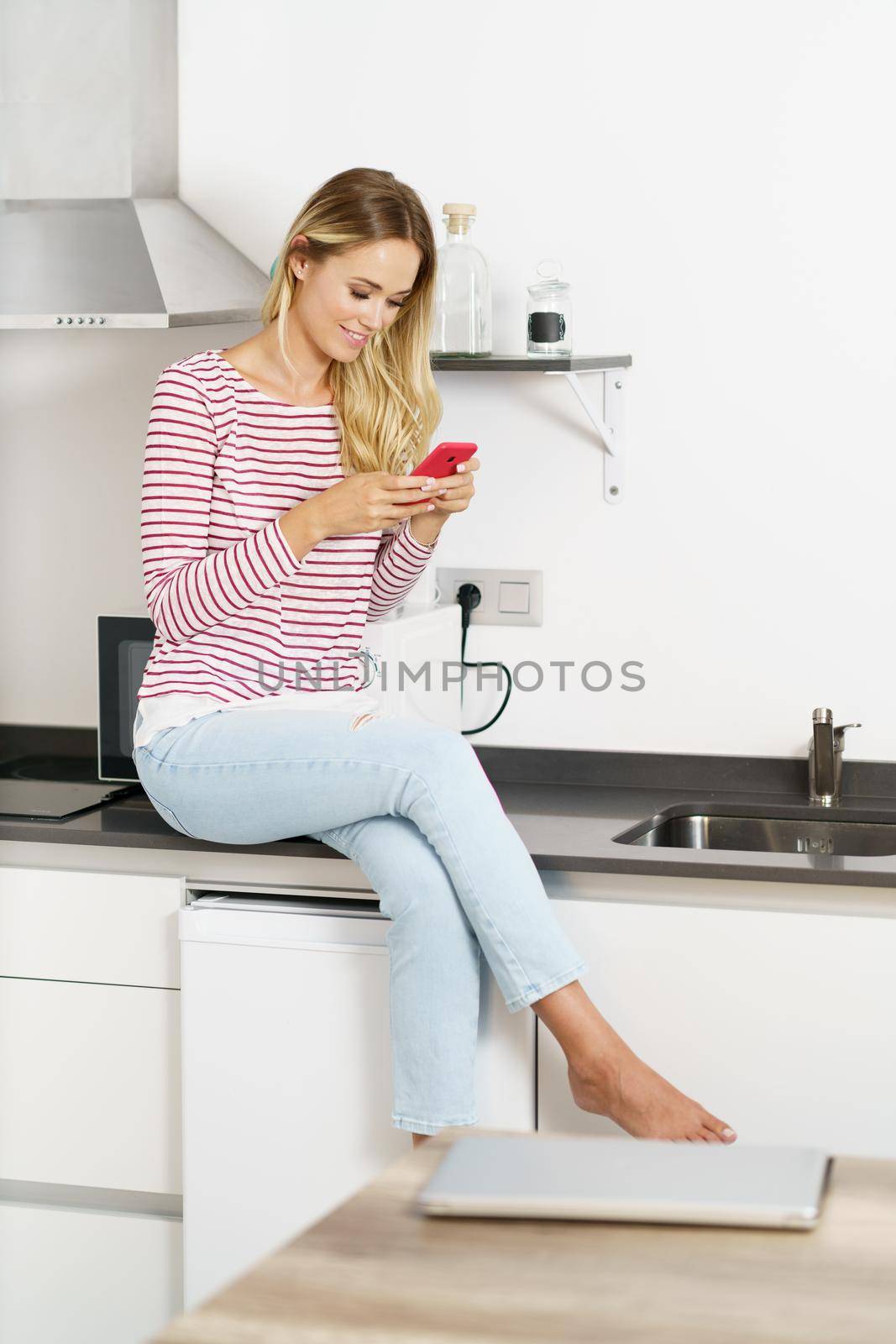 Happy woman using her smartphone sitting in the kitchen at home