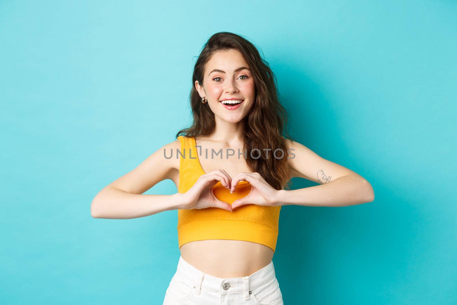 Summer holidays and emotions concept. Young romantic girl showing I love you heart gesture and smiling, standing over blue background. Copy space