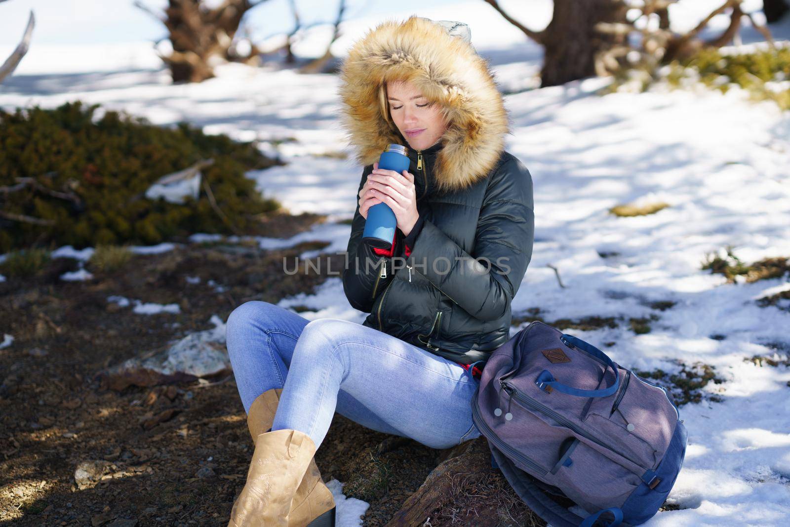 Woman drinking something hot from a metal thermos bottle sitting on a rock in the snowy mountains. by javiindy