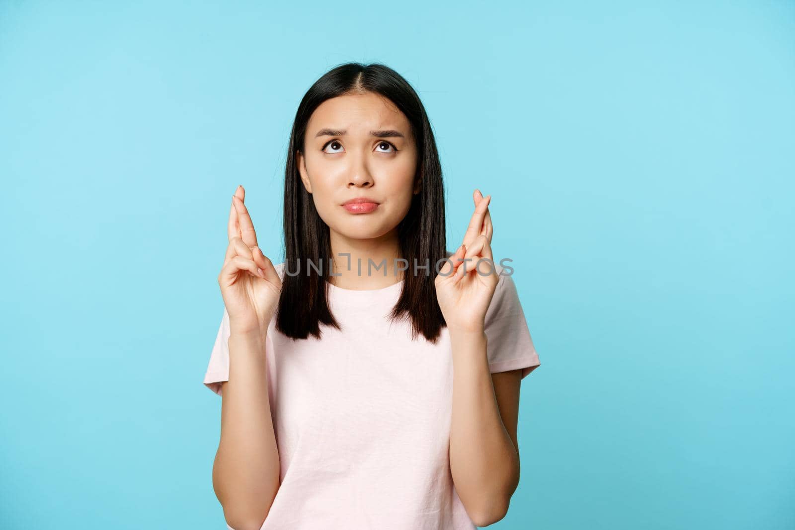 Worried asian girl looking up hopeful and nervous, cross fingers for good luck, making wish, praying, standing in t-shirt over blue background.