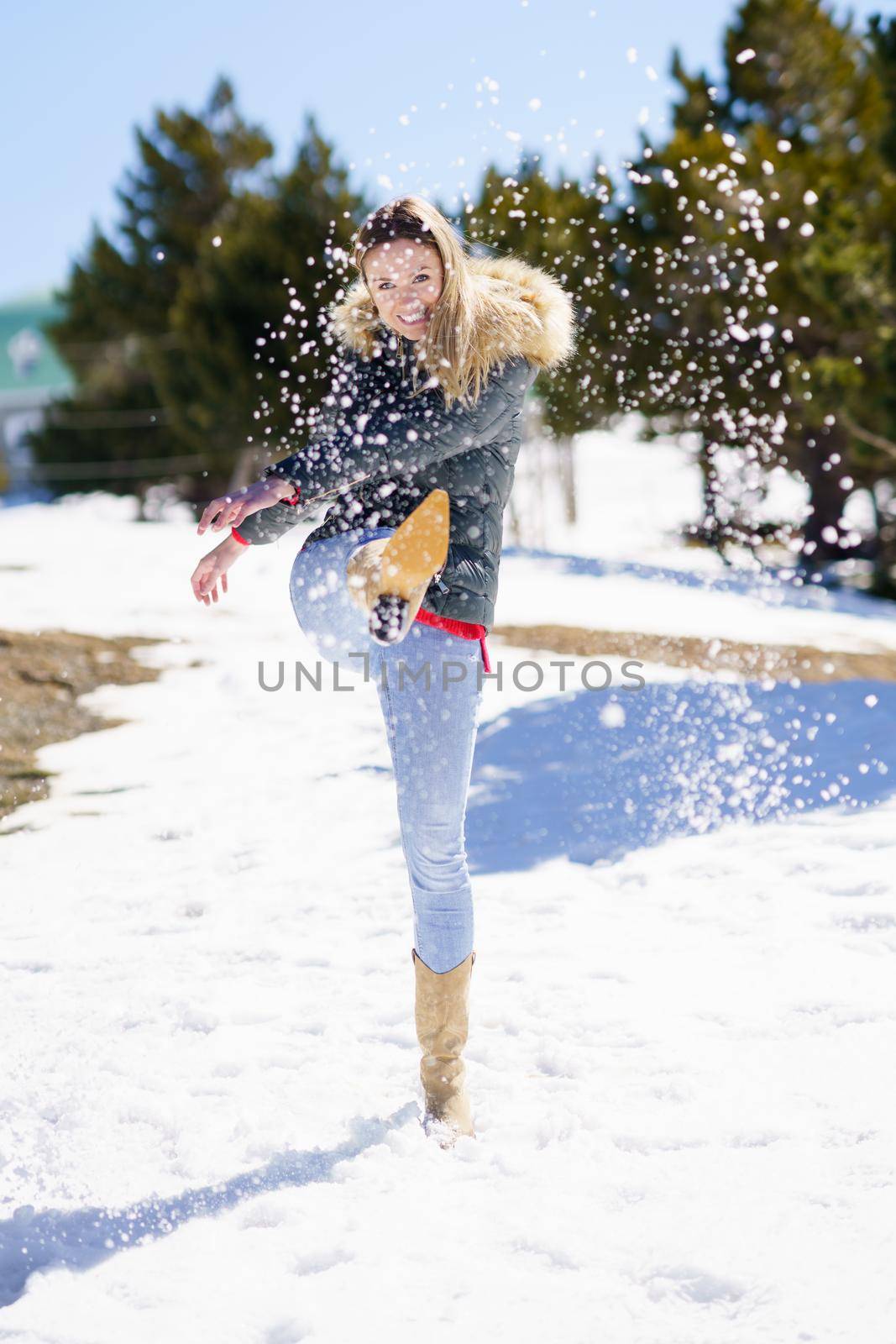 Young happy woman kicking snow in a snow-covered forest in the mountains in Sierra Nevada, Granada, Spain. Female wearing winter clothes playing with snow.