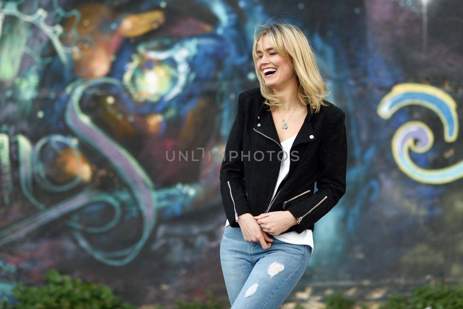 Funny blonde woman laughing in urban background. Young girl wearing black zipper jacket and blue jeans trousers standing in the street. Pretty female with straight hair hairstyle and blue eyes.