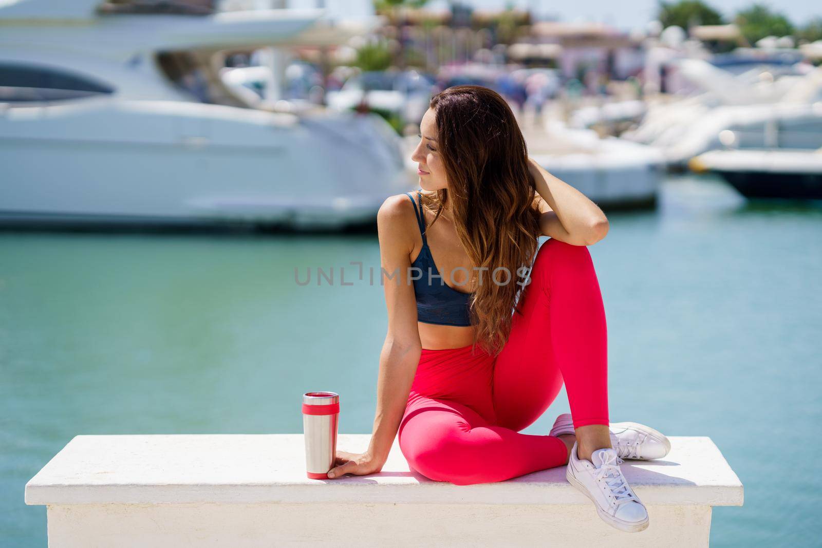 Fitness girl in red sportswear outfit training resting to hydrate after exercise on waterfront harbour.