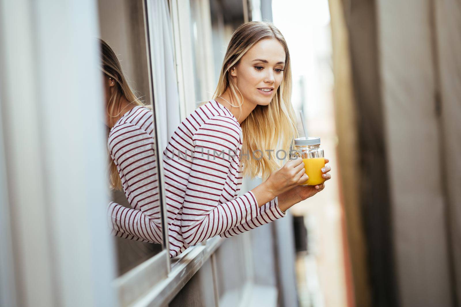 Young woman drinking a glass of natural orange juice, leaning out the window of her home. by javiindy