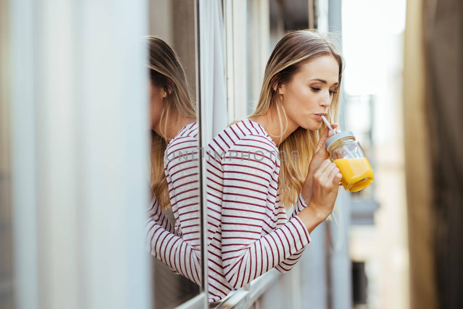 Young woman drinking a glass of natural orange juice, leaning out the window of her home. by javiindy