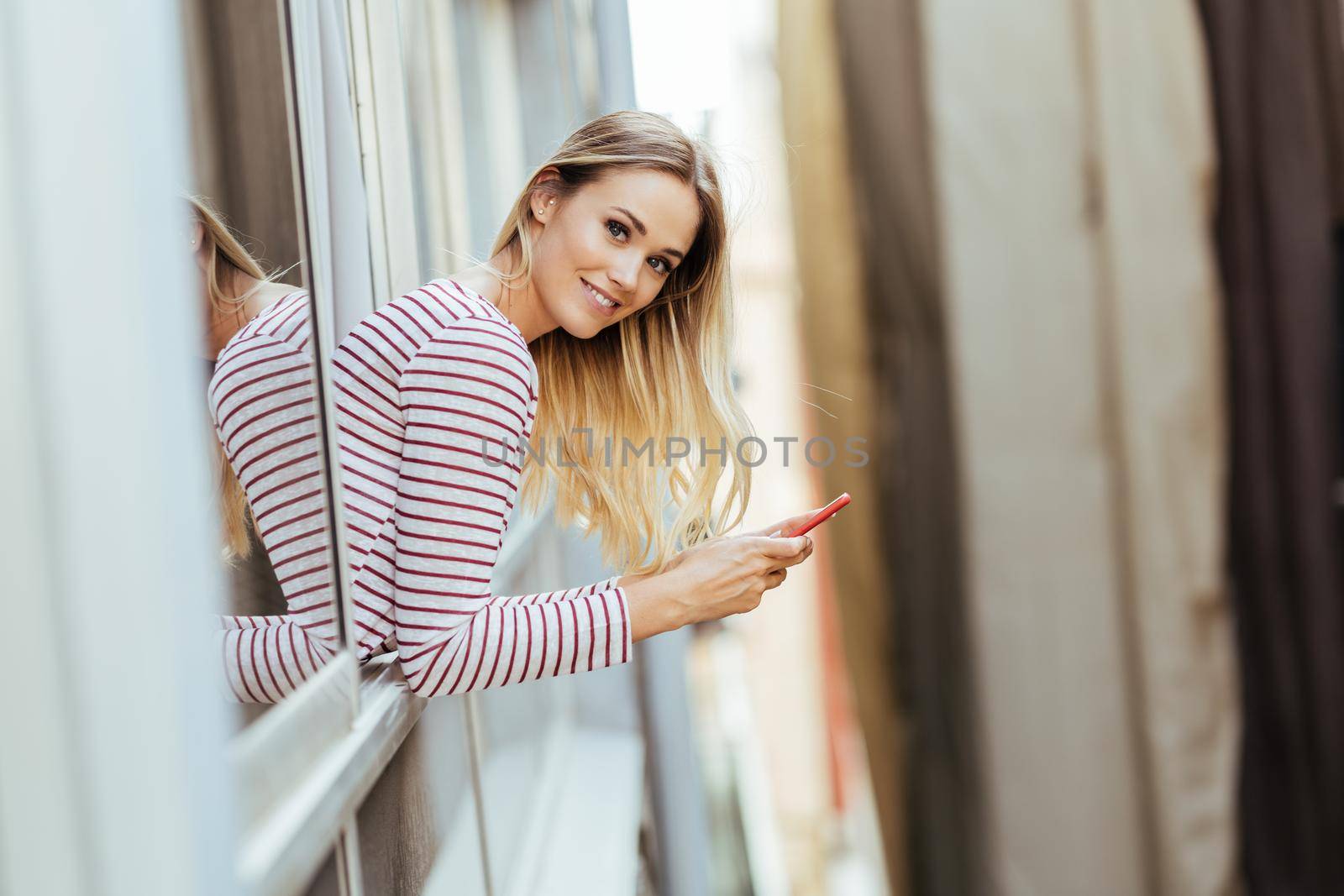 Young woman leaning out of her house window using a smartphone. by javiindy