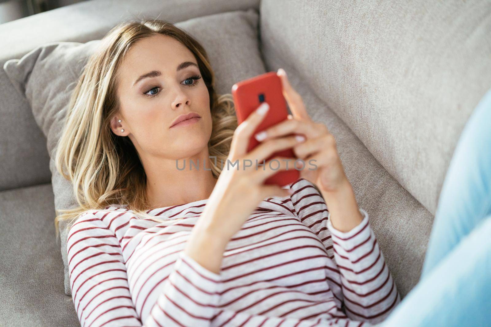 Young blonde woman using her smartphone lying on the sofa. by javiindy