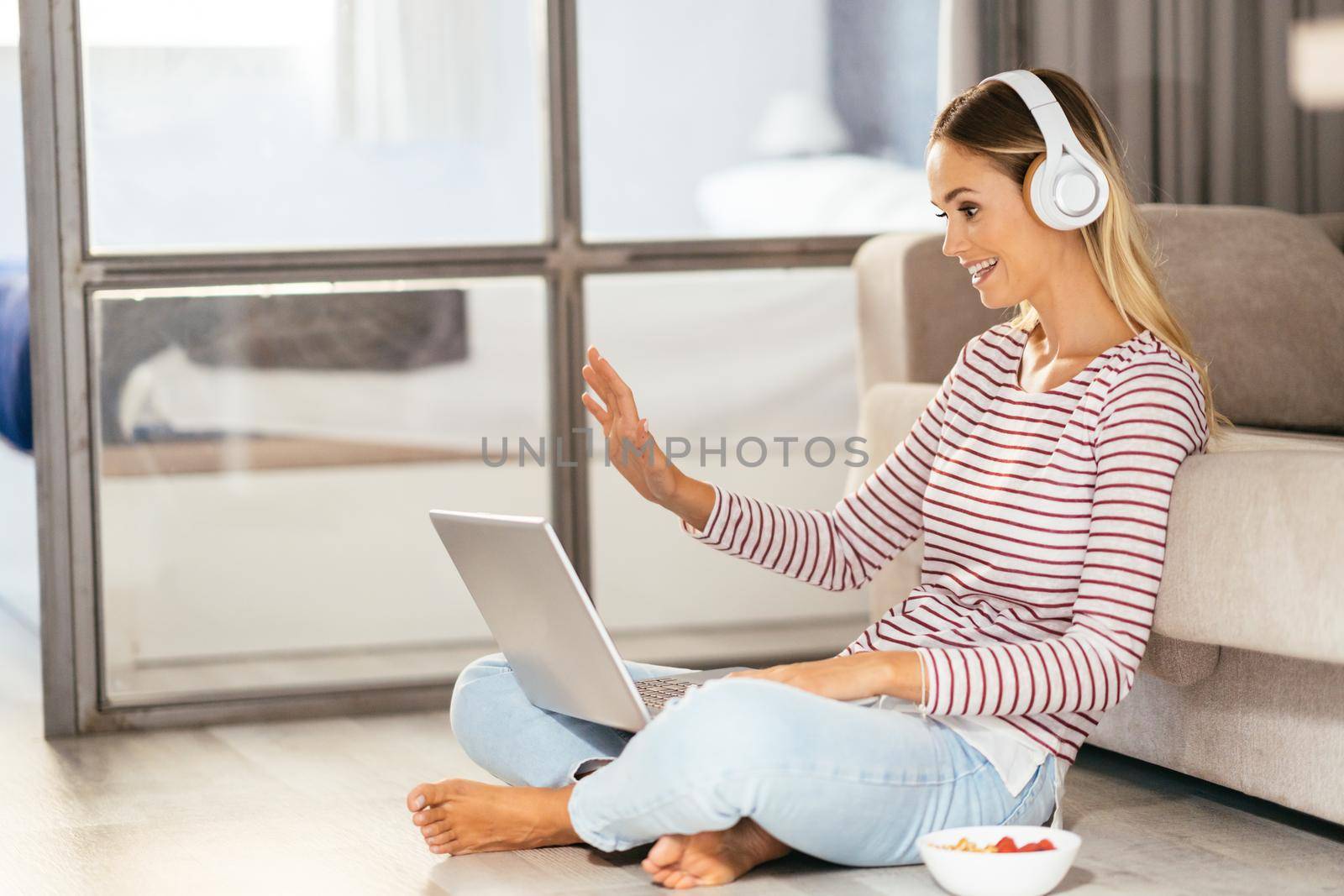 Smiling young woman with headphones and laptop videoconferencing on the sofa at home.